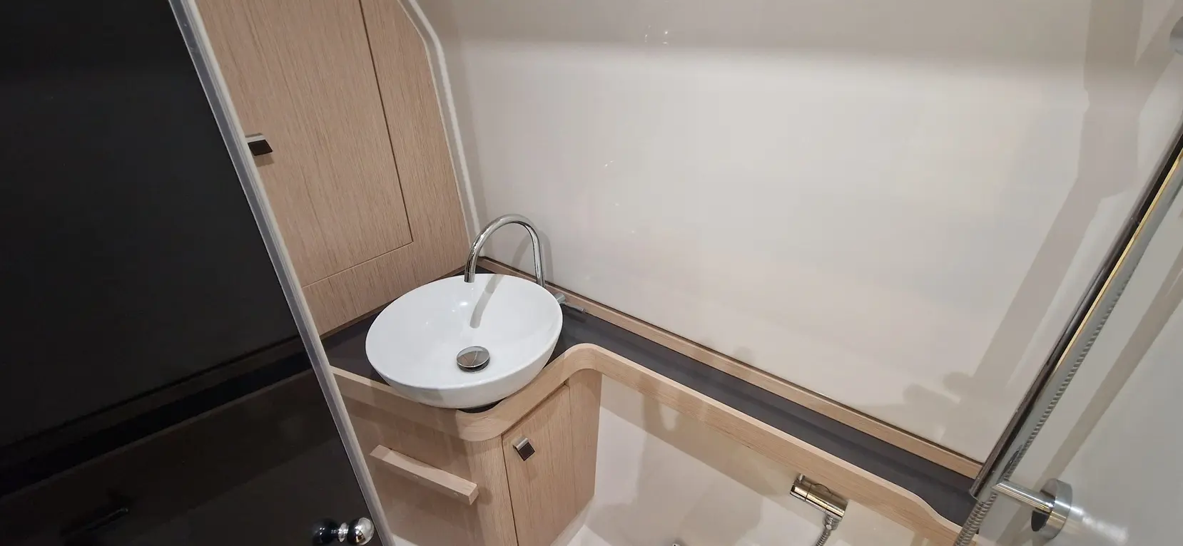 Modern bathroom featuring a white oval sink, chrome tap, and wooden cabinetry against a plain wall.
